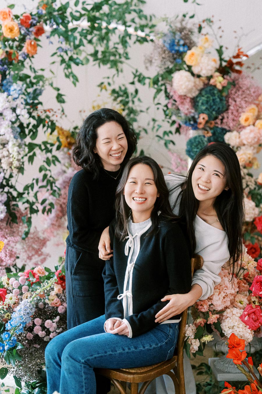 Three women smiling and posing together in front of a vibrant, colorful floral arrangement, embodying happiness and friendship.