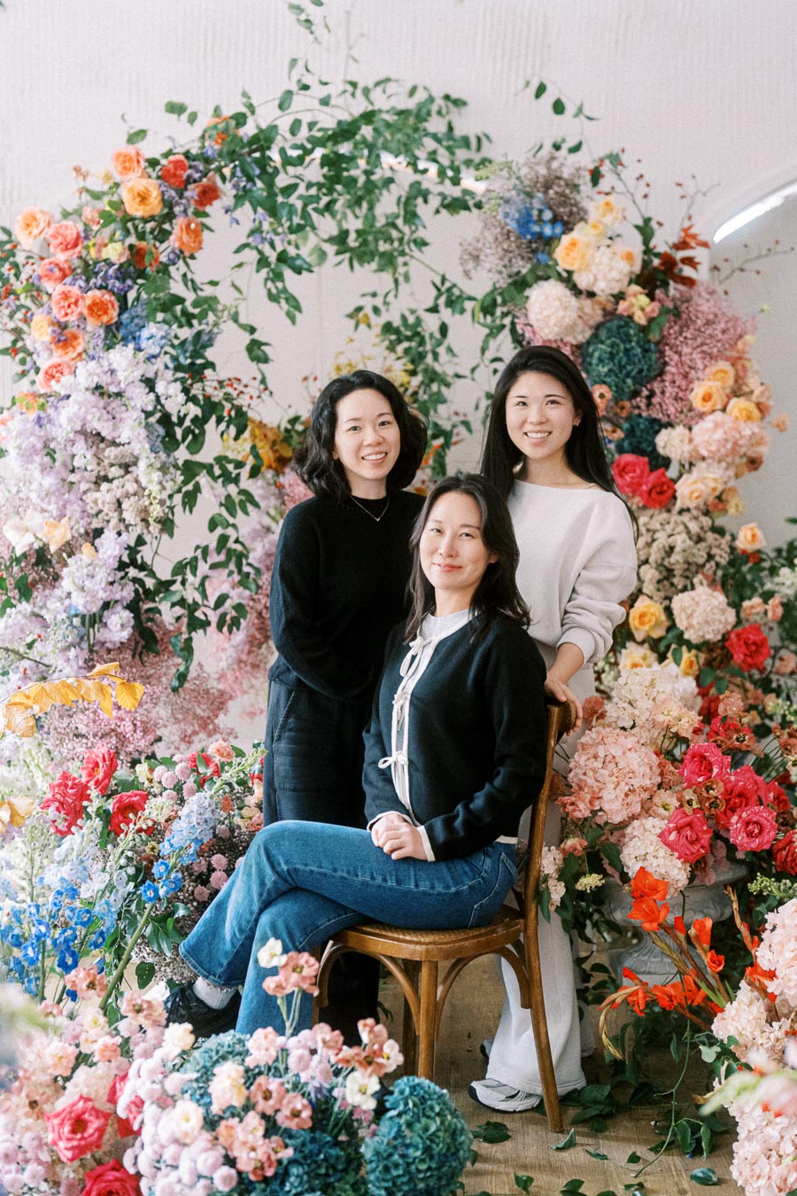 Three women surrounded by vibrant floral arrangements, showcasing colorful roses, hydrangeas, and greenery, creating a lush and artistic backdrop.