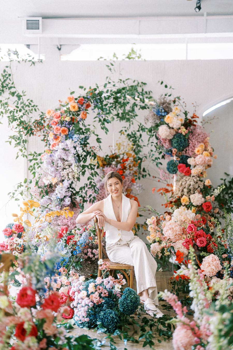 A woman sitting on a wooden chair surrounded by a vibrant array of colorful flowers and greenery, creating a lush floral backdrop indoors.