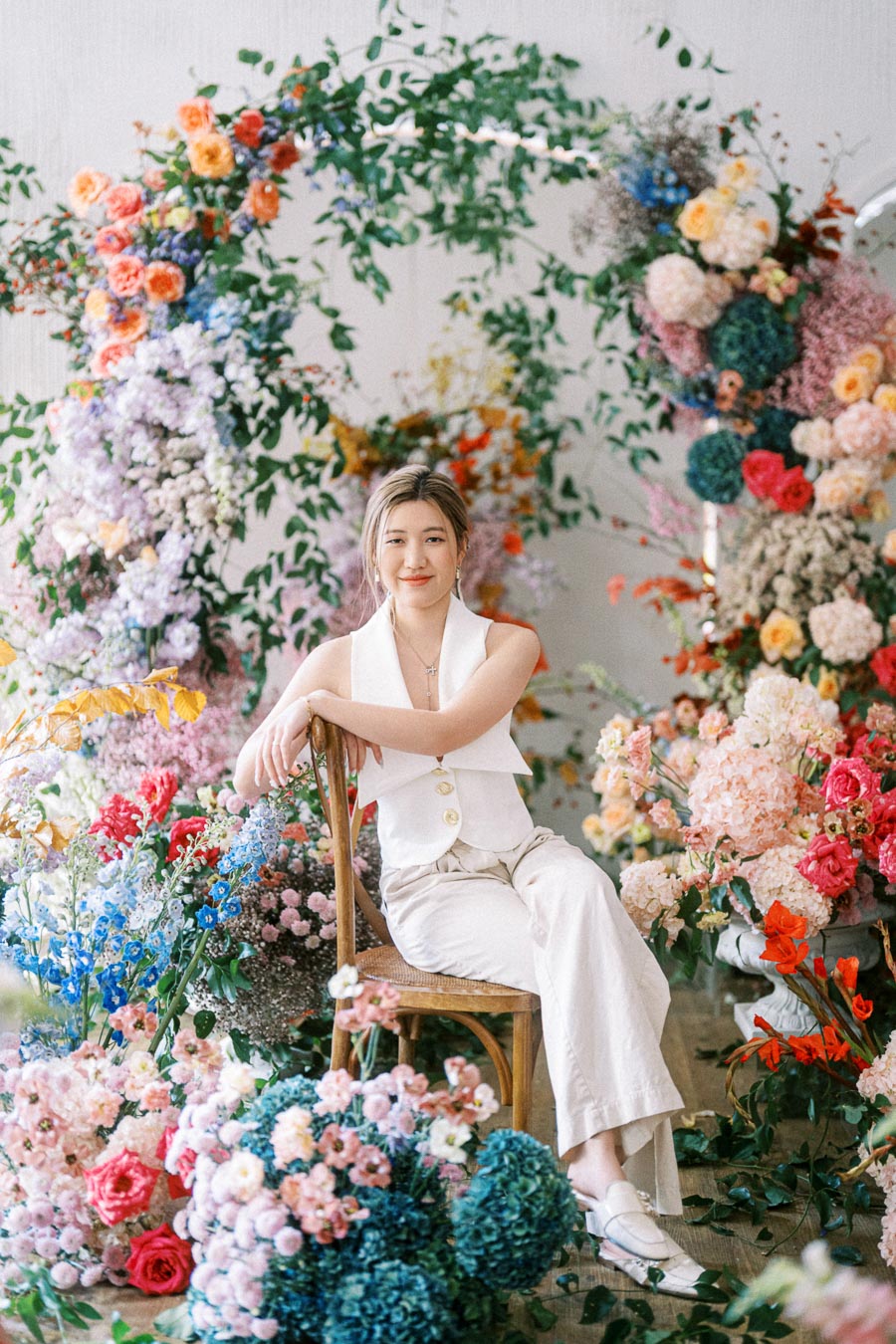 A person seated on a wooden chair surrounded by vibrant, colorful floral arrangements, wearing a white outfit. The scene is set indoors with an elegant and artistic backdrop, featuring an array of flowers including roses, hydrangeas, and greenery.