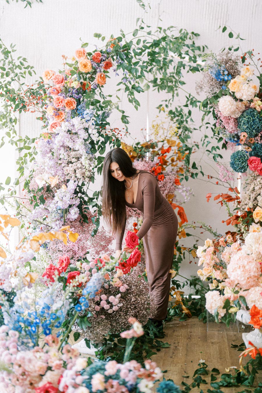 A woman in a brown dress arranging a vibrant display of colorful flowers and greenery in a whimsical botanical setting.
