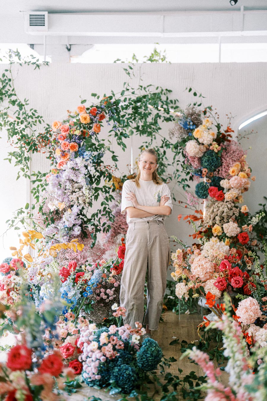 A person standing in front of a vibrant flower arrangement featuring a variety of colorful blooms and lush green foliage, showcasing a floral design display in a well-lit interior space.