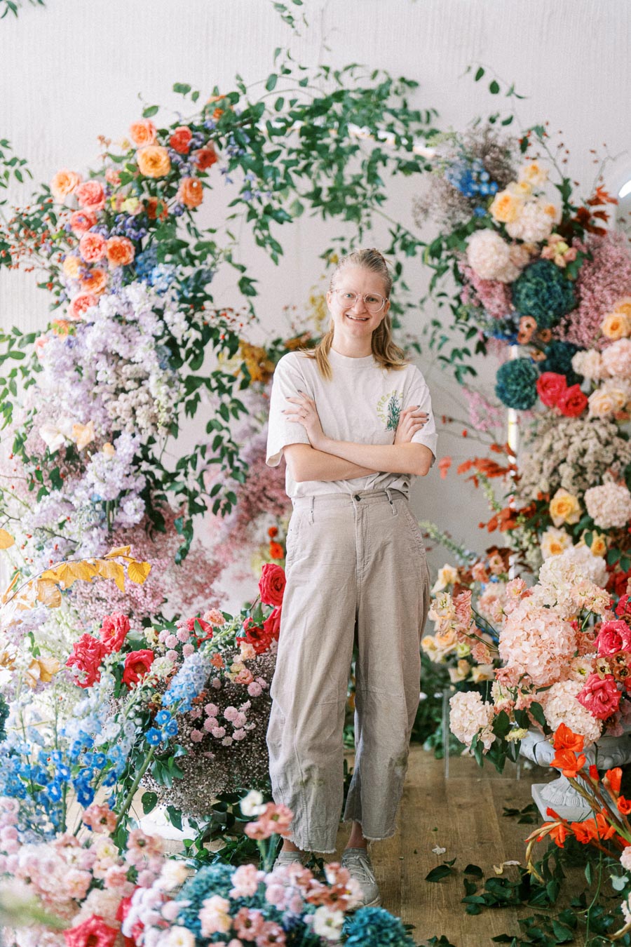A smiling person stands surrounded by a vibrant array of colorful flowers in an indoor setting, dressed casually with arms crossed, conveying a cheerful and creative atmosphere.