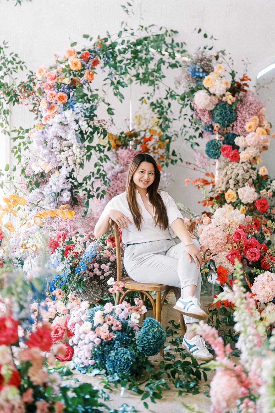 A person sitting on a chair surrounded by a vibrant and colorful floral arrangement, featuring a variety of blooms in shades of pink, red, blue, and orange, against a neutral background.