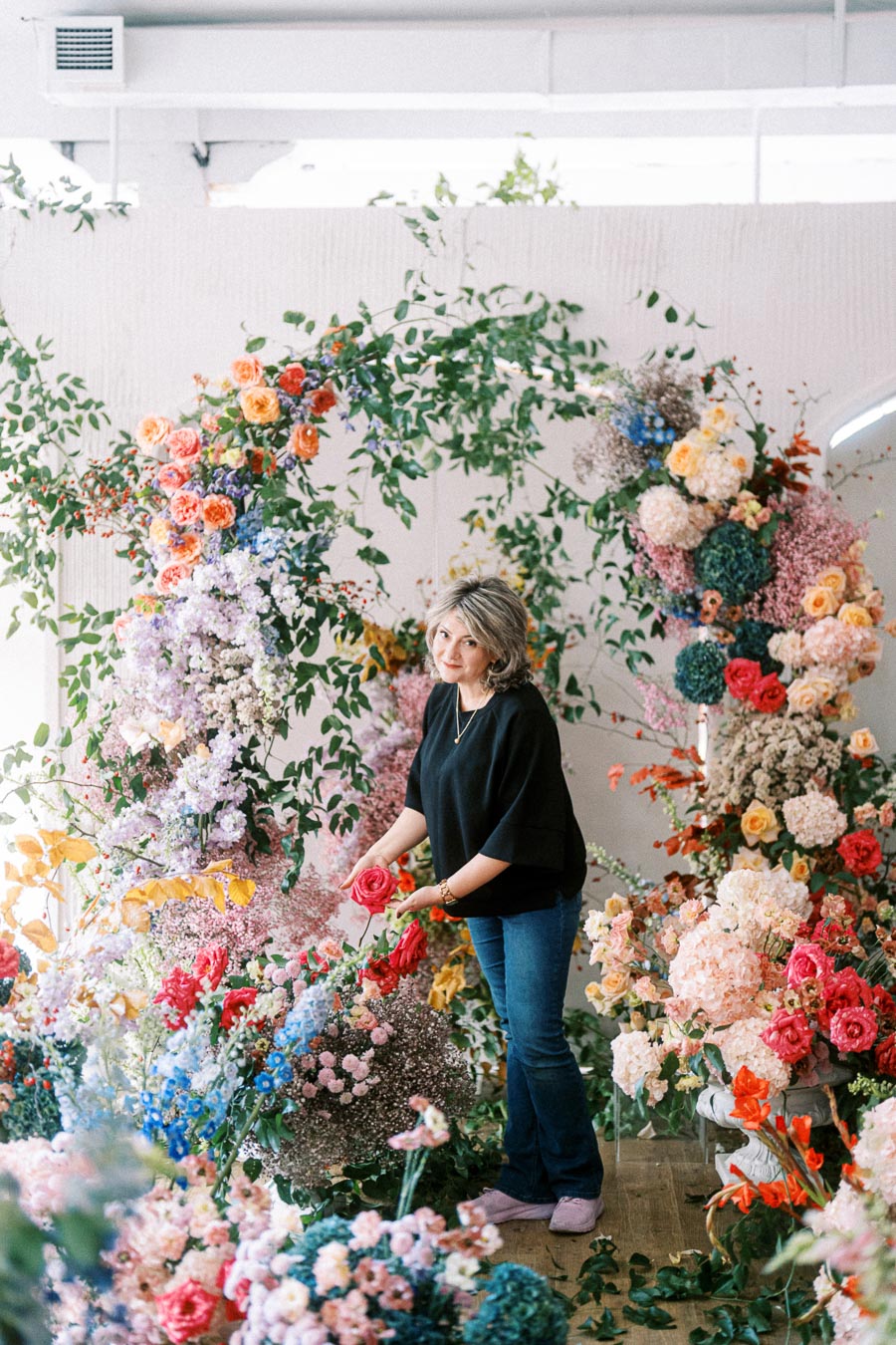 A florist arranging a vibrant floral arch with a variety of colorful flowers, including roses and hydrangeas, in a bright studio setting.