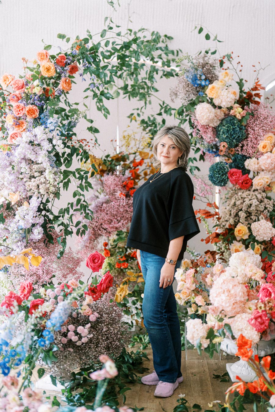A woman standing amidst a vibrant floral archway, surrounded by colorful roses, hydrangeas, and greenery, dressed in a black top and blue jeans.