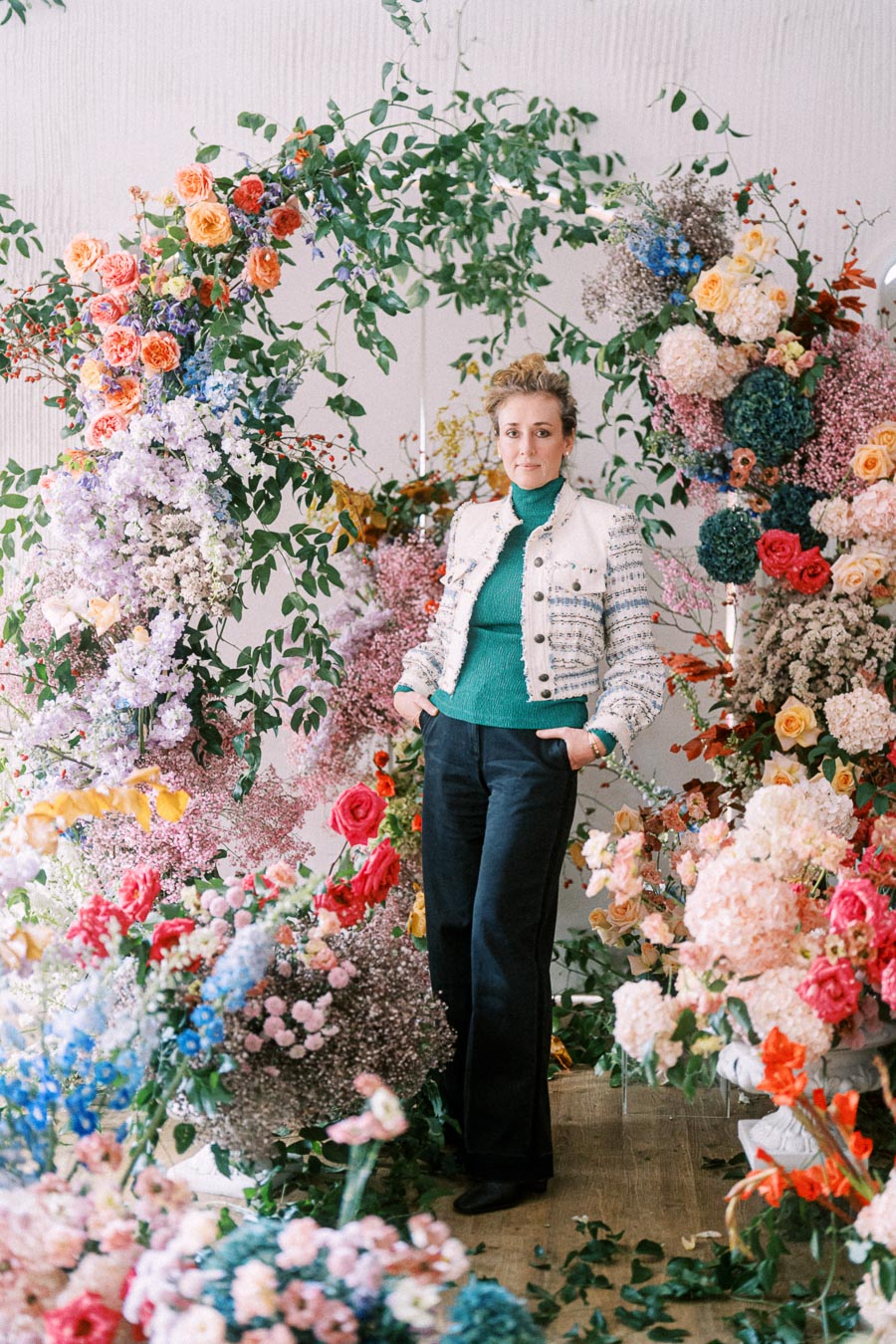 A person in stylish attire stands amidst an elaborate floral arrangement featuring vibrant roses, hydrangeas, and lush greenery, creating a visually striking display of colorful blooms.