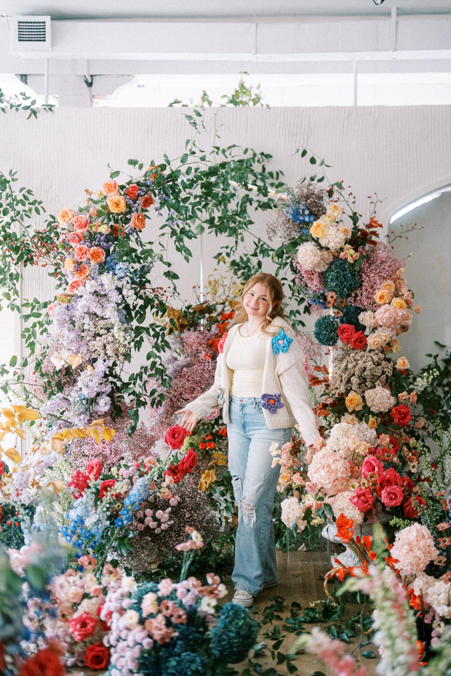 A person standing in a lush indoor garden surrounded by vibrant, colorful flowers and green leaves, creating a whimsical and enchanting atmosphere.