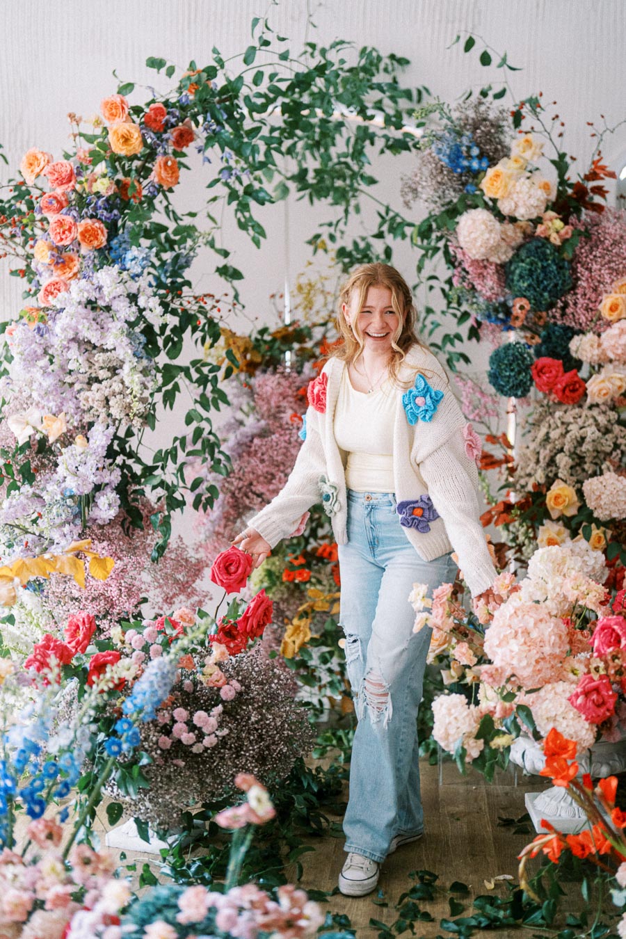 Smiling person standing amidst a vibrant indoor floral display, surrounded by colorful roses, hydrangeas, and mixed greenery, evoking a joyful and serene atmosphere.