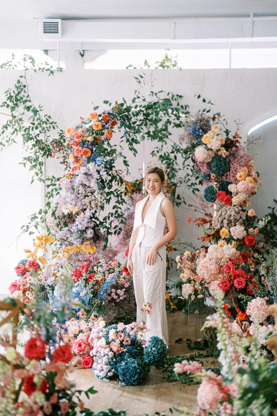 Person in white outfit standing amidst a vibrant floral display with a variety of colorful flowers and greenery forming an artistic backdrop. Perfect for a spring-themed event or garden inspiration.