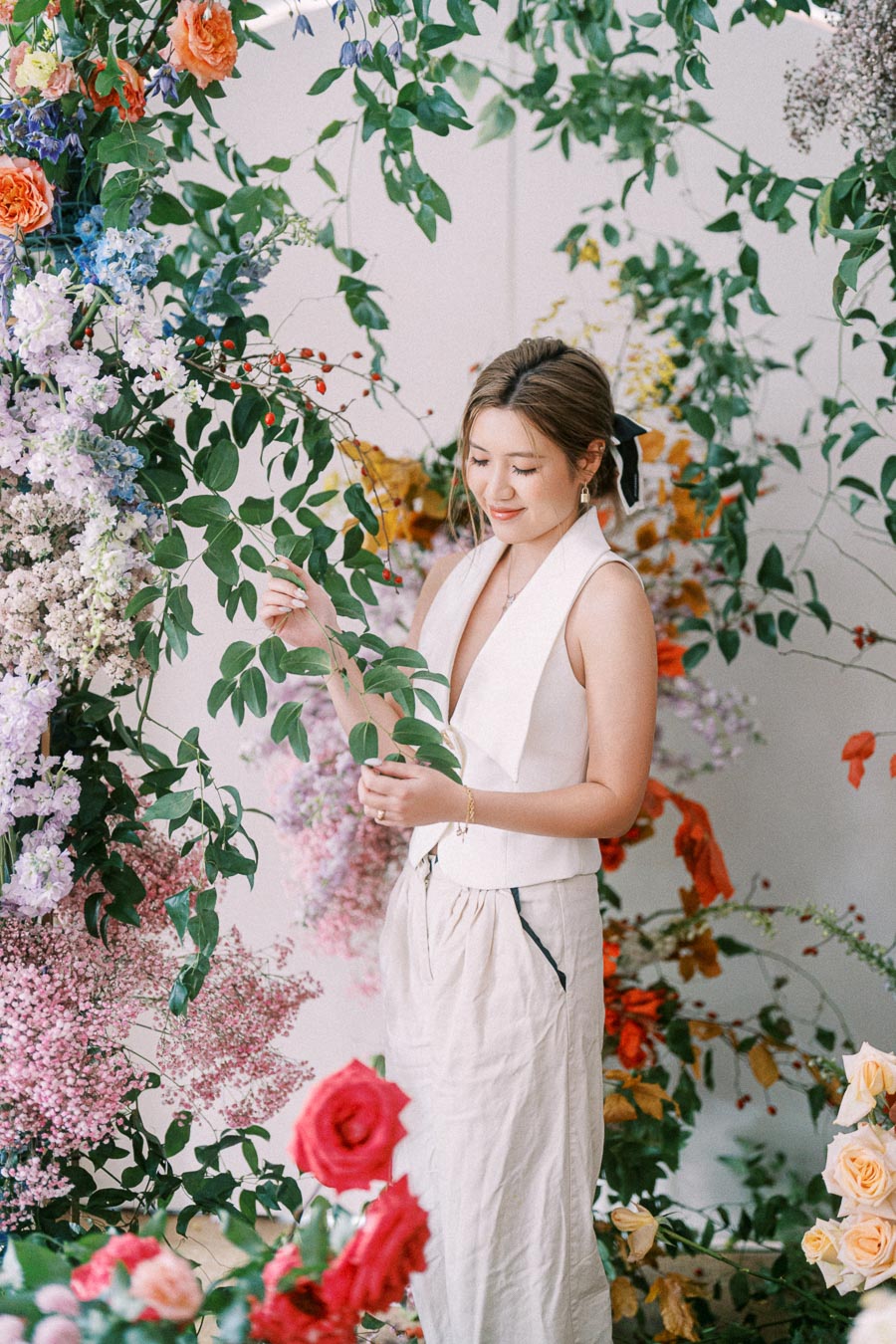 A woman in a white dress standing among vibrant flowers and green leaves, surrounded by colorful floral arrangements in shades of red, pink, and orange.