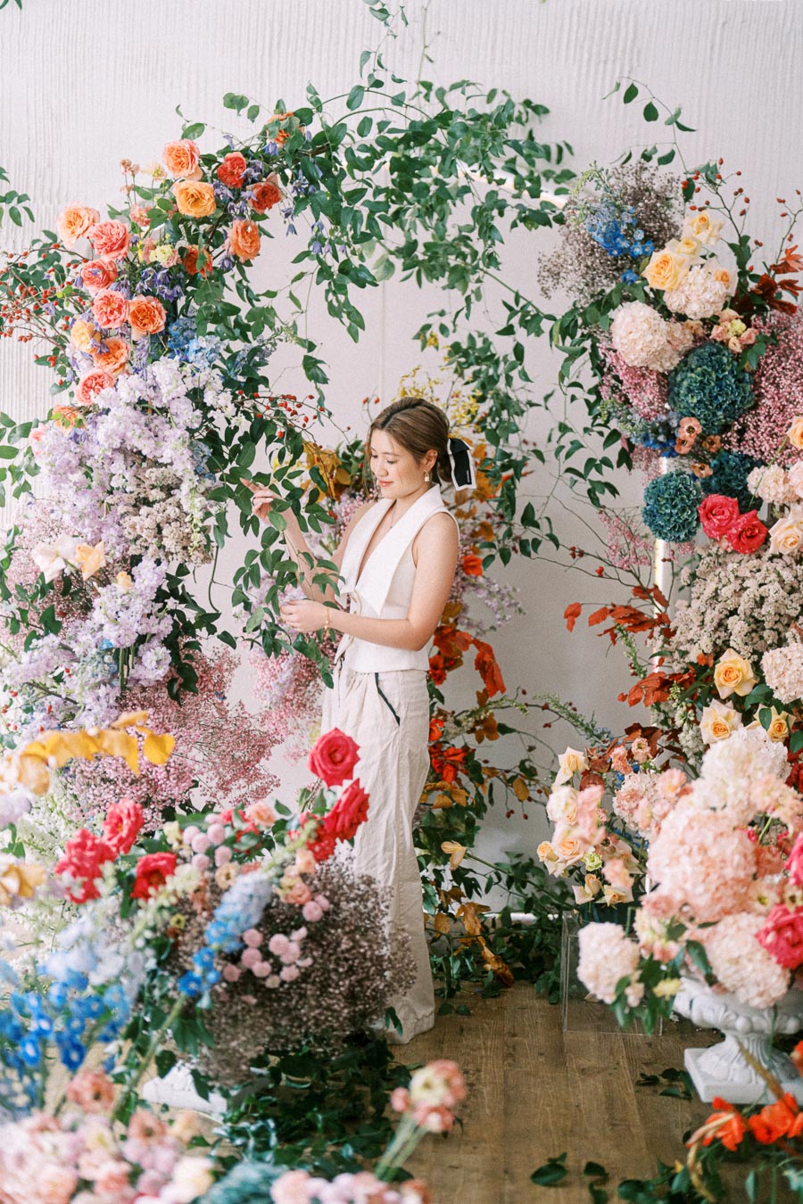 A woman arranging a vibrant display of colorful flowers and greenery, creating an elegant and artistic floral arrangement in a bright indoor setting.