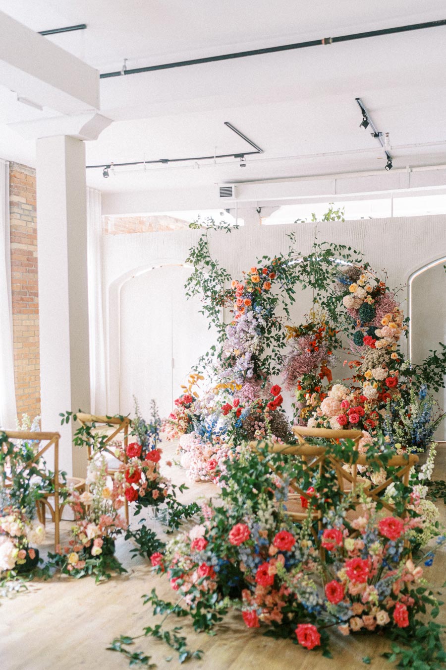 Elegant indoor floral installation featuring colorful flowers and greenery arranged around wooden chairs, set against a bright, open space with modern design elements. Perfect for weddings or events.