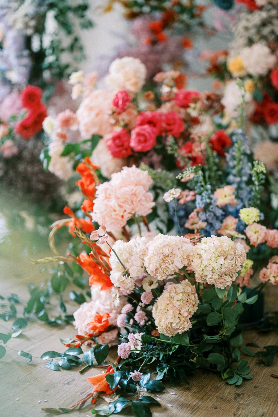 A vibrant assortment of colorful flowers, including soft pink hydrangeas, vivid red roses, and orange gladiolus, arranged beautifully on a wooden surface.