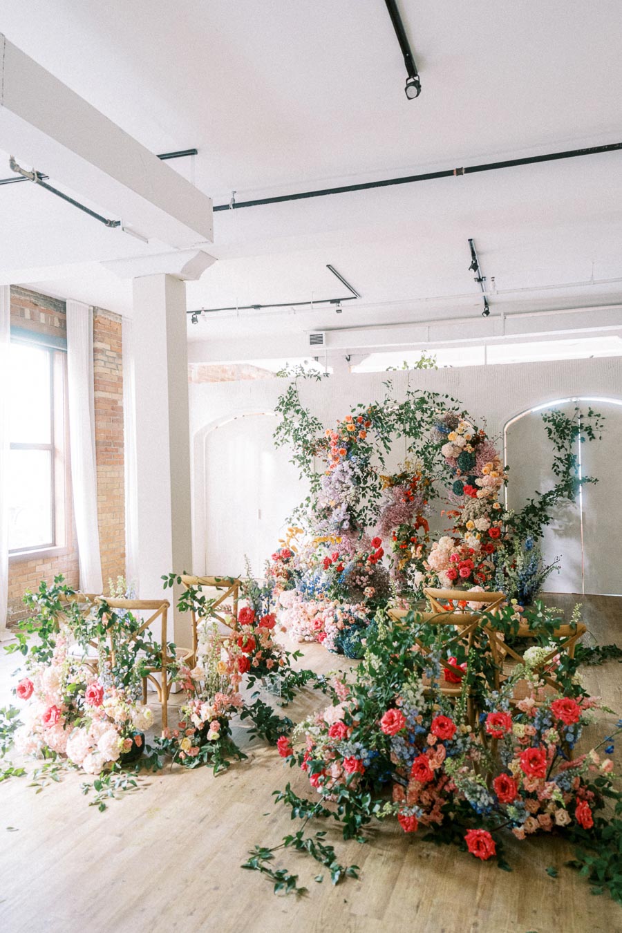 Bright and airy event space with a stunning floral installation featuring vibrant roses, peonies, and greenery cascading over wooden chairs, creating a whimsical and romantic setting.