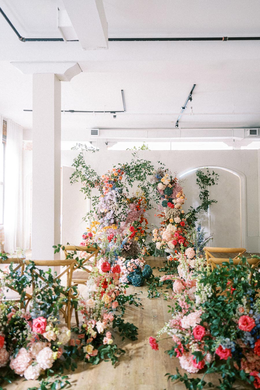 Luxurious indoor wedding ceremony setup with a vibrant floral archway and elegant garden-style arrangements featuring roses, hydrangeas, and greenery on wooden flooring.