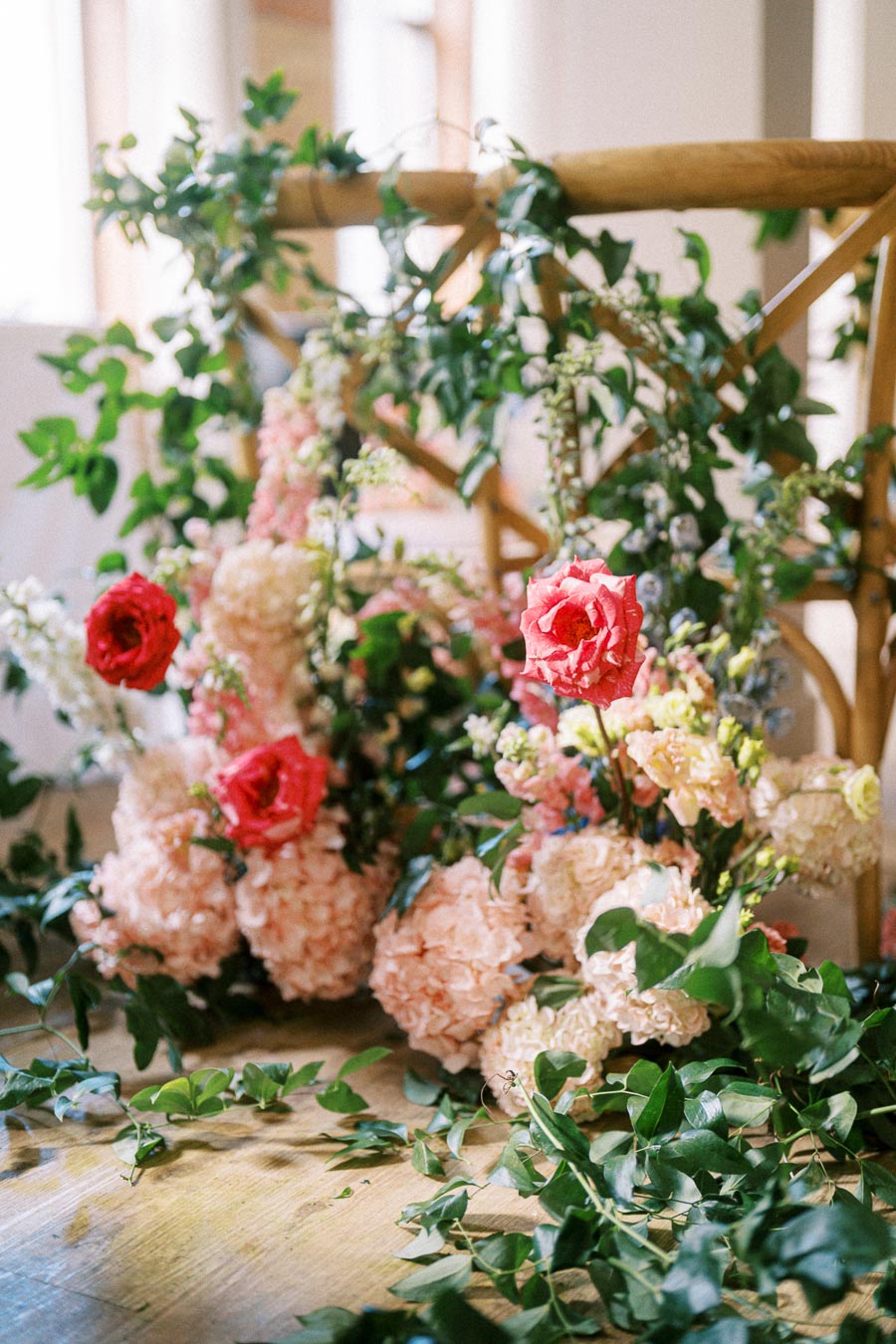Beautiful floral arrangement featuring vibrant red and pink roses, surrounded by lush greenery and delicate pink hydrangeas on a wooden chair, evoking a fresh and elegant garden setting.
