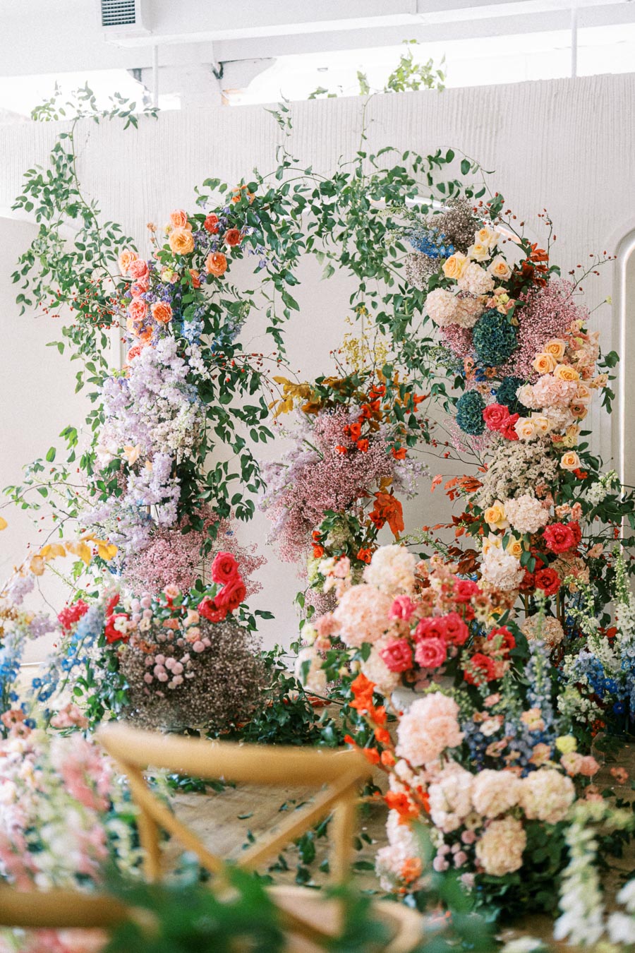 A vibrant floral arrangement featuring a diverse mix of colorful flowers, including roses, baby's breath, and greenery, creating an elegant arch display in a bright, indoor setting.