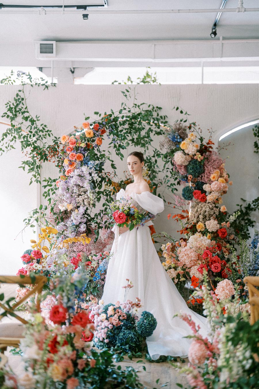 Bride in elegant white gown surrounded by vibrant floral arrangements in a modern indoor setting.