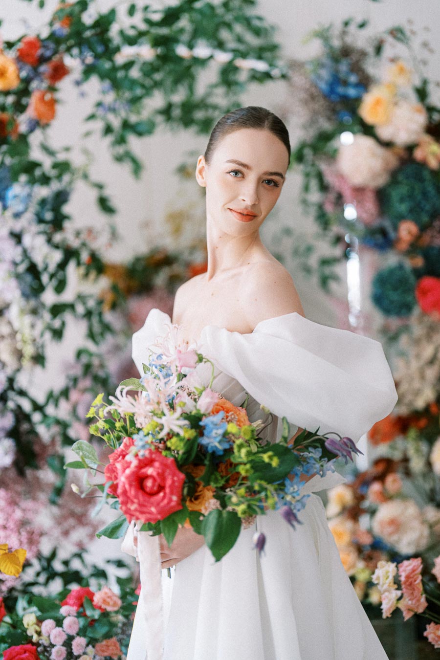 A bride in an elegant white dress holding a vibrant bouquet of colorful flowers, standing amidst a lush floral backdrop.