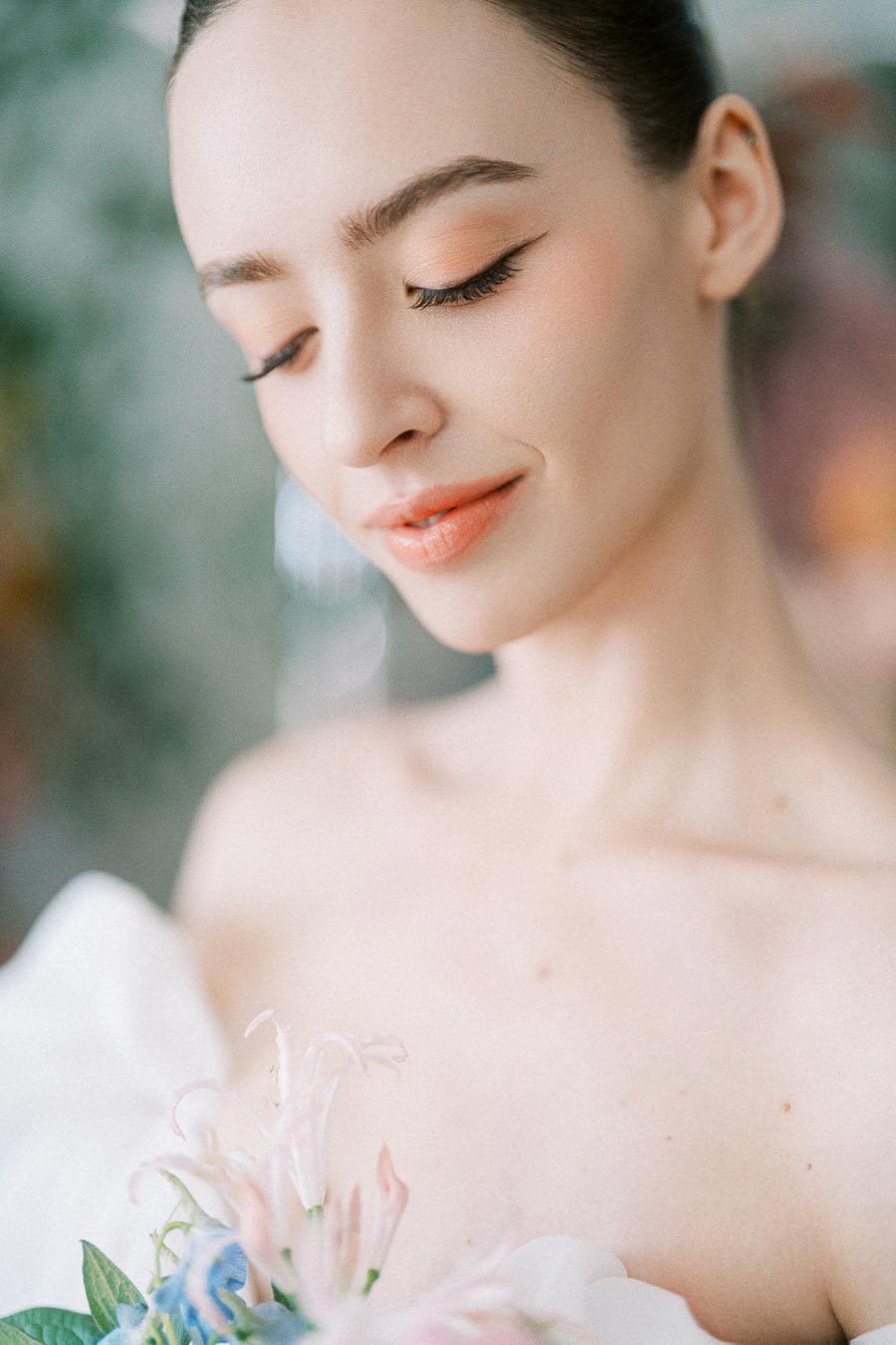 Beautiful bride with soft makeup smiling while holding a bouquet of delicate pink and blue flowers against a blurred background.