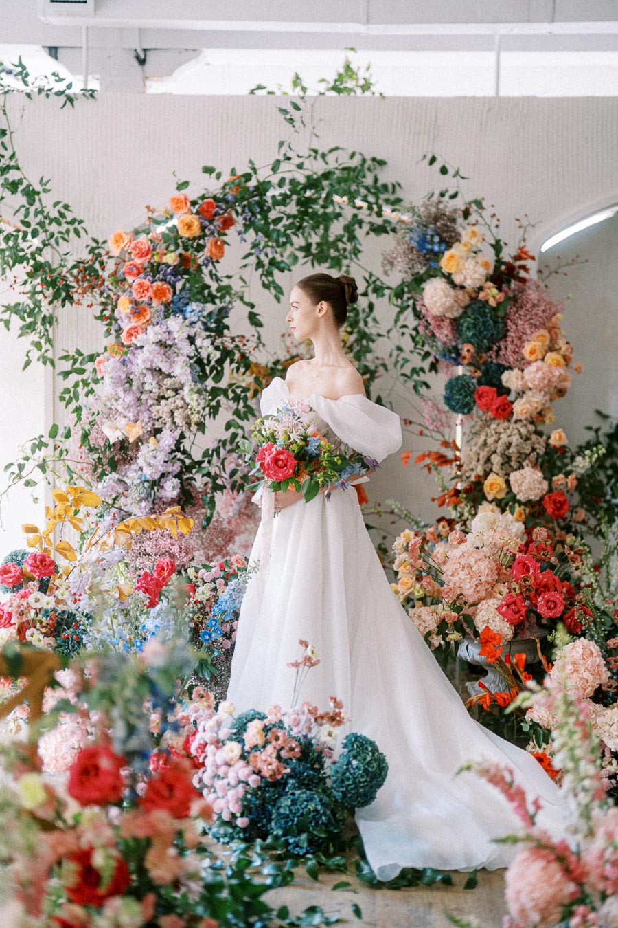 A bride in an off-shoulder white gown stands surrounded by a vibrant floral arch featuring colorful blooms like roses, hydrangeas, and peonies, creating a romantic and whimsical wedding setting.