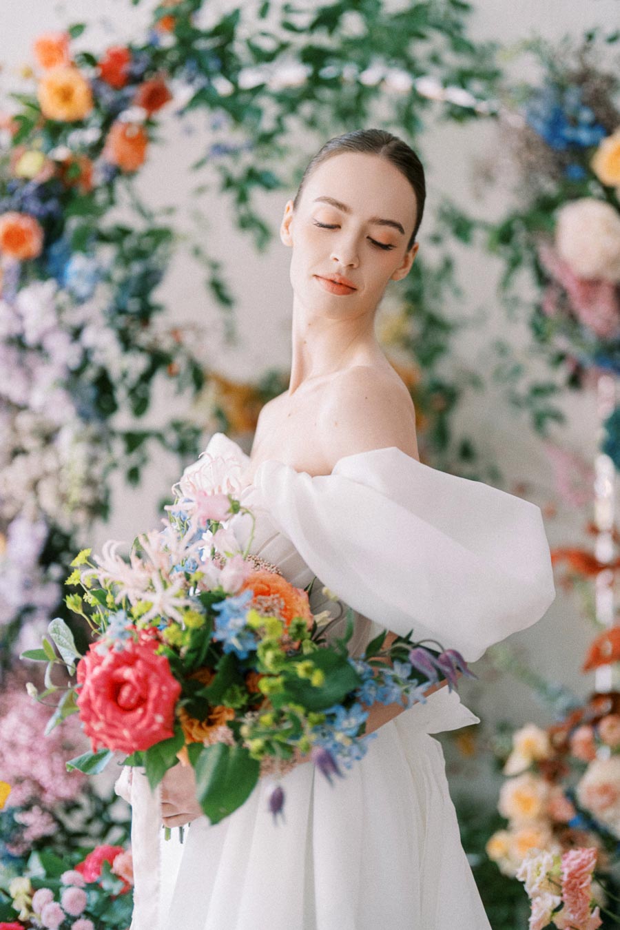 Bride in a white off-shoulder dress holding a colorful bouquet, surrounded by vibrant floral arrangements.