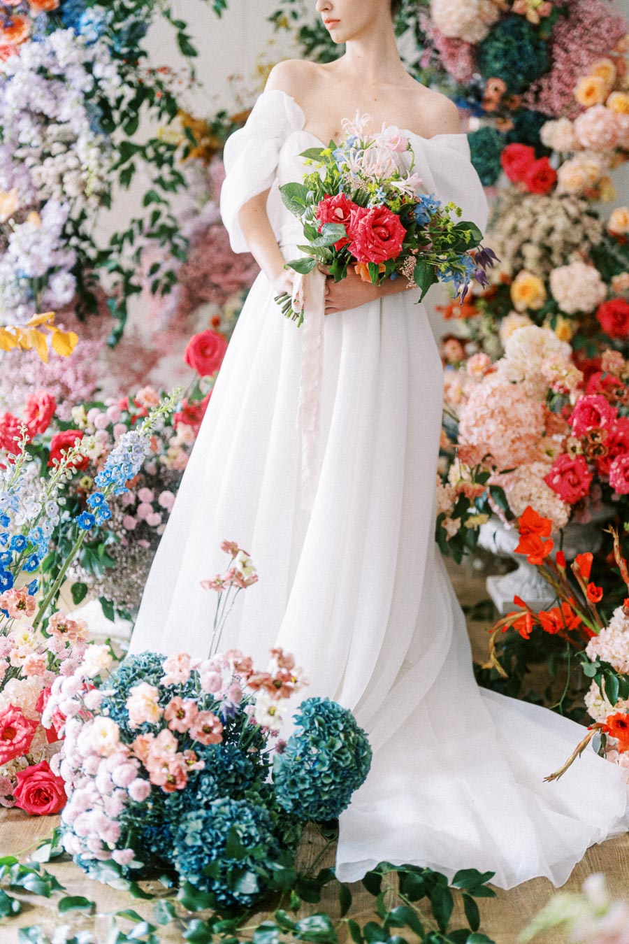 A bride in an elegant off-the-shoulder white wedding dress holding a vibrant bouquet, surrounded by a lush floral backdrop with colorful blossoms and greenery.