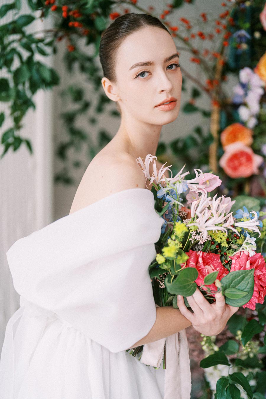 A woman in an off-the-shoulder white dress holding a vibrant bouquet of flowers, including pink, blue, and yellow blossoms, set against a lush, leafy background.