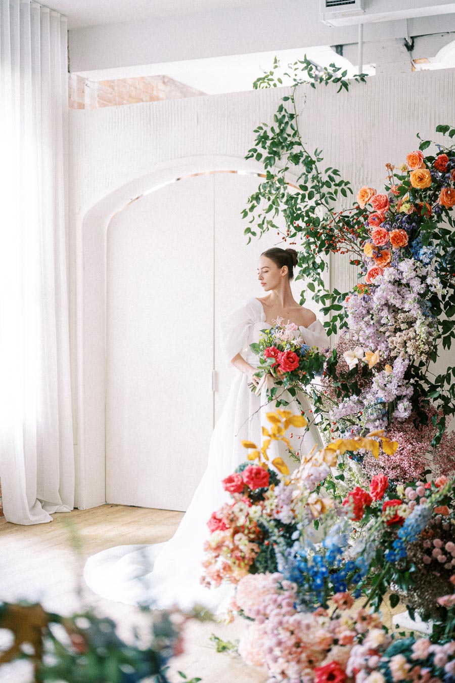 A bride in an elegant white wedding dress stands next to a vibrant floral arrangement featuring colorful flowers, including roses and lilacs, inside a light-filled room with white curtains.