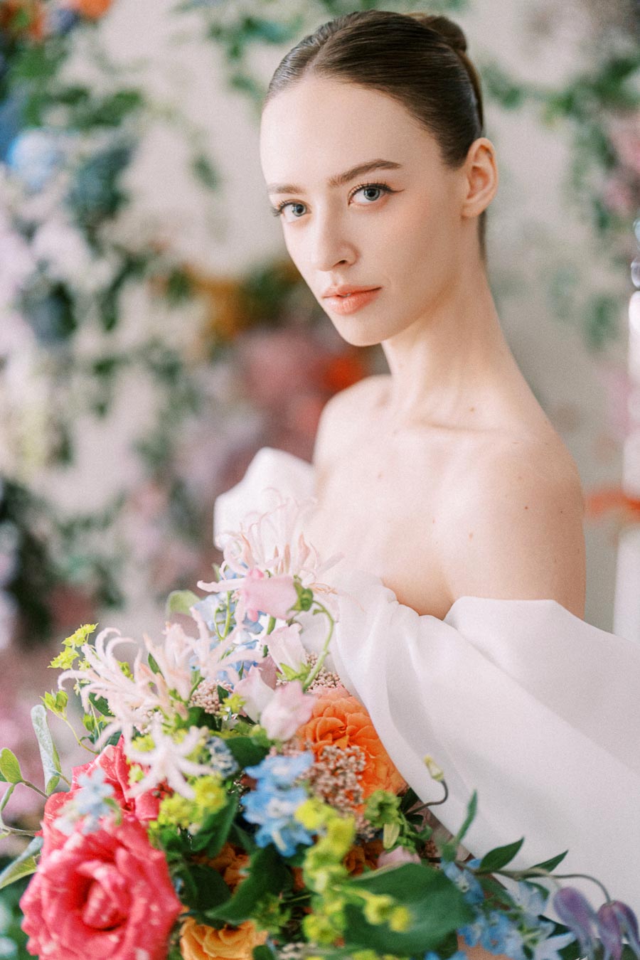 Elegant woman holding vibrant bouquet of colorful flowers, wearing a white dress, blurred floral background.