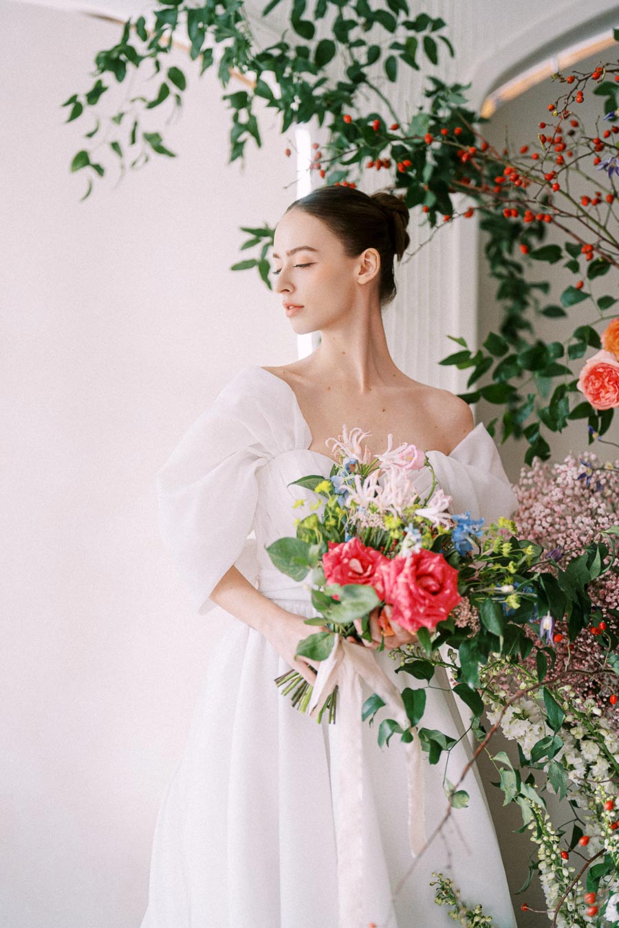 Bride in elegant white wedding dress holding a vibrant floral bouquet, surrounded by lush greenery and colorful flowers.