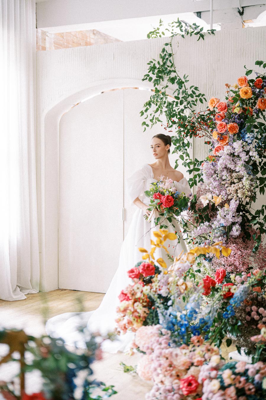 A bride in a white dress stands elegantly beside a vibrant floral arrangement, featuring a variety of colorful flowers, in a bright, well-lit room with white curtains and a modern archway.