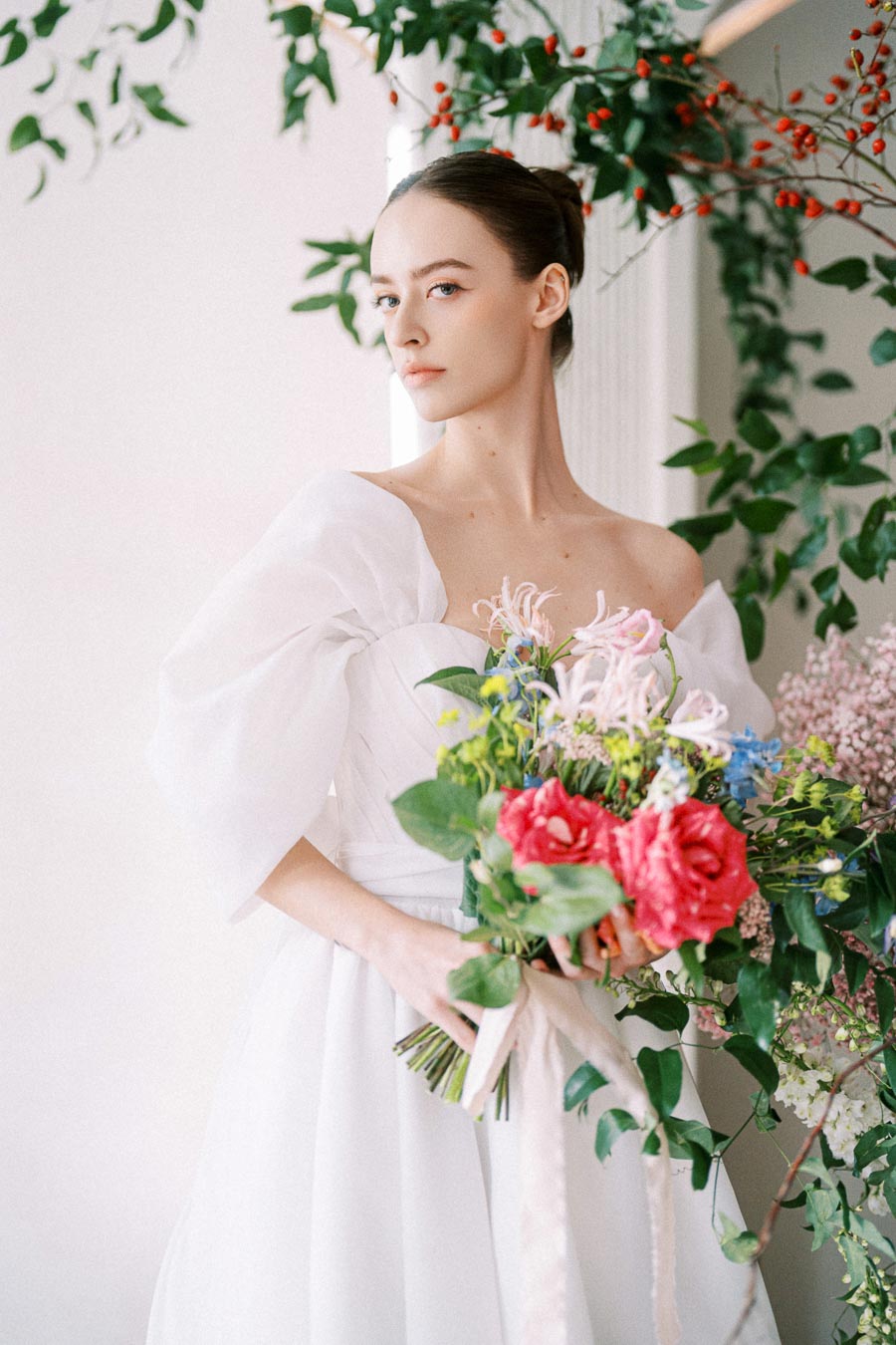 A bride in an elegant white off-shoulder wedding dress holds a vibrant bouquet of flowers, surrounded by lush green foliage and red berries, creating a romantic and picturesque scene.