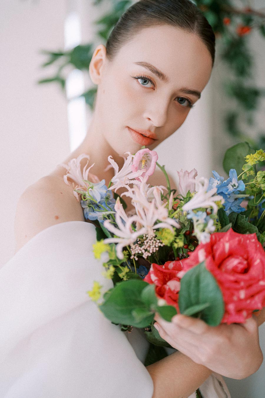 A woman holding a colorful bouquet of flowers including pink, red, and blue blossoms, surrounded by green foliage, with a soft-focus background.