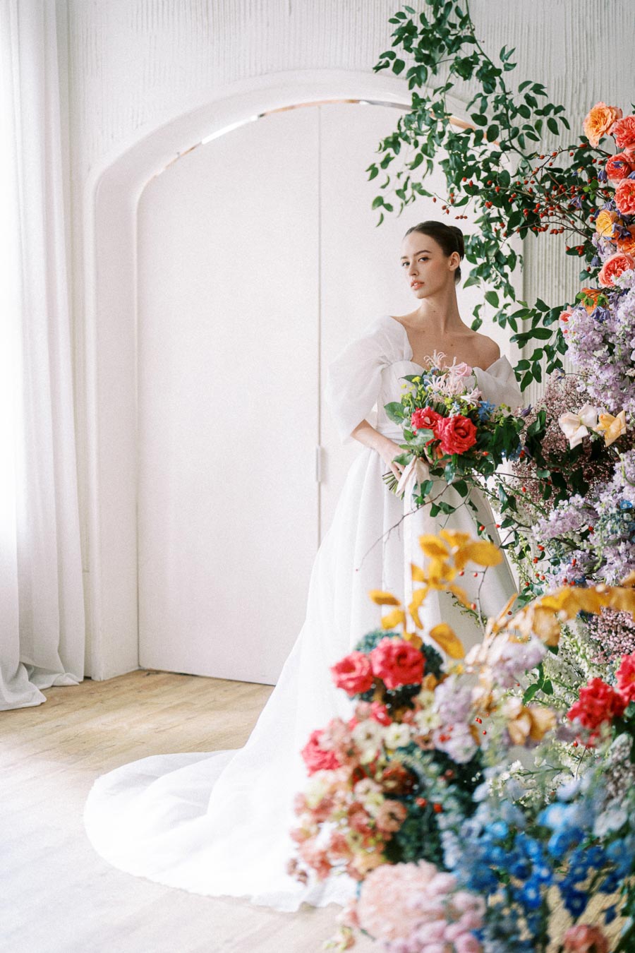 A bride in a white wedding gown holding a vibrant bouquet of flowers stands gracefully in a beautifully decorated room with colorful floral arrangements and lush greenery, creating an elegant and romantic atmosphere.