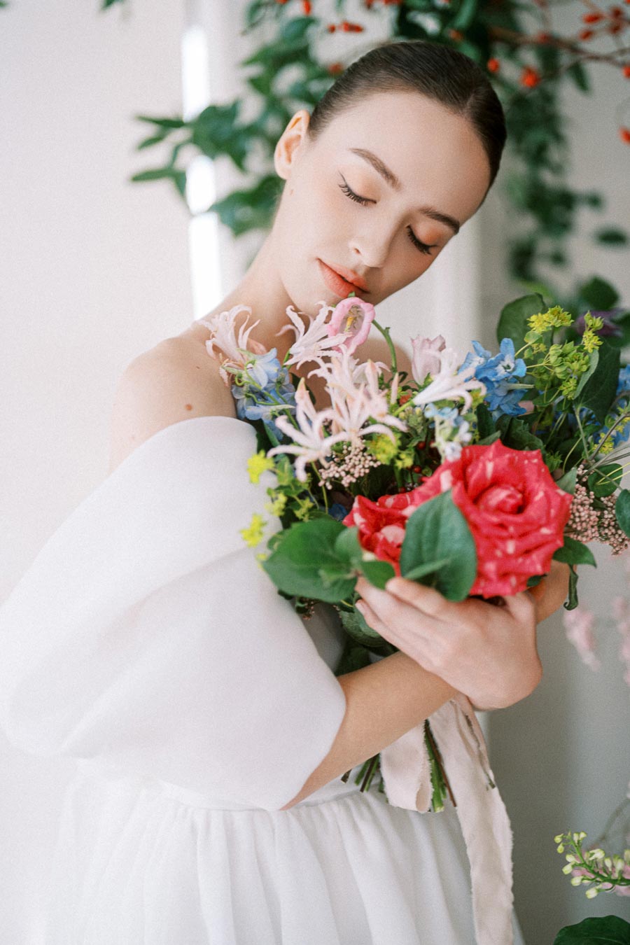 A woman in a white dress gently holds a colorful bouquet of flowers, including pink lilies, blue blossoms, and a vibrant red rose, set against a soft-focus background of greenery.