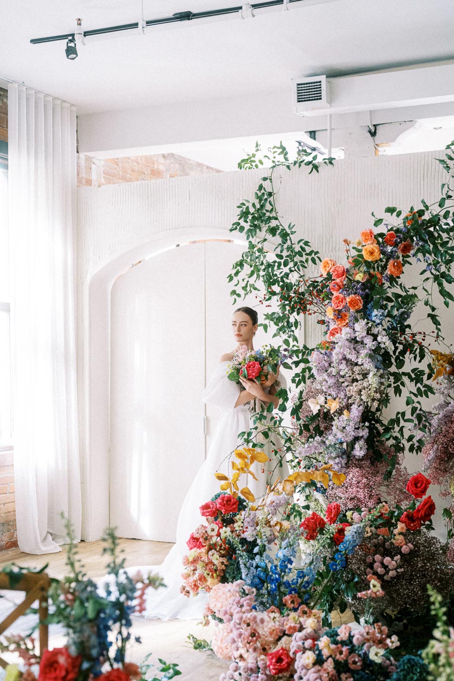 A bride in a white gown holding a bouquet stands among an elaborate floral display featuring vibrant roses and greenery in a bright, airy room.