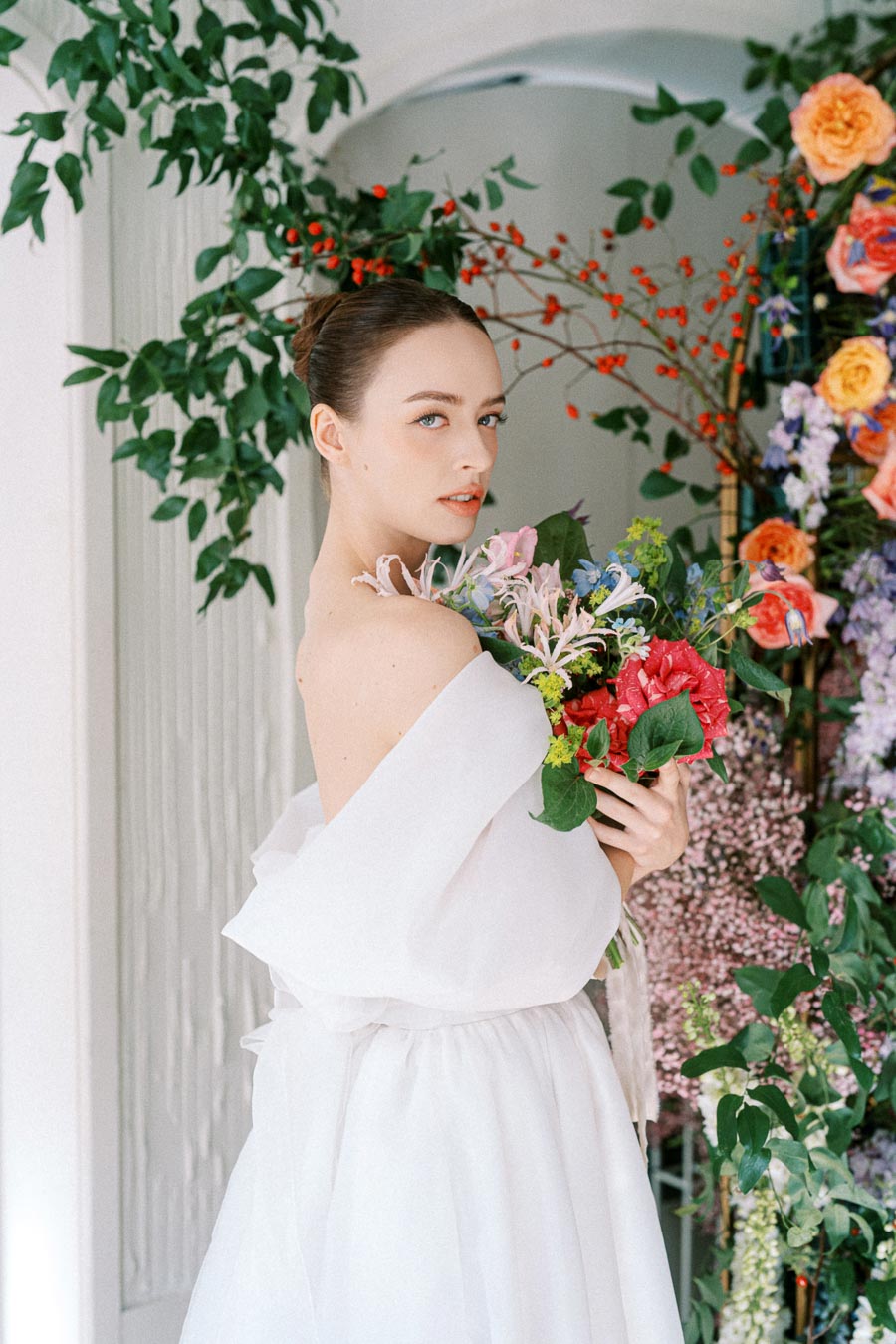 Elegant bride in a white gown holding a vibrant bouquet of flowers, surrounded by lush greenery and colorful blossoms, in a serene indoor setting.