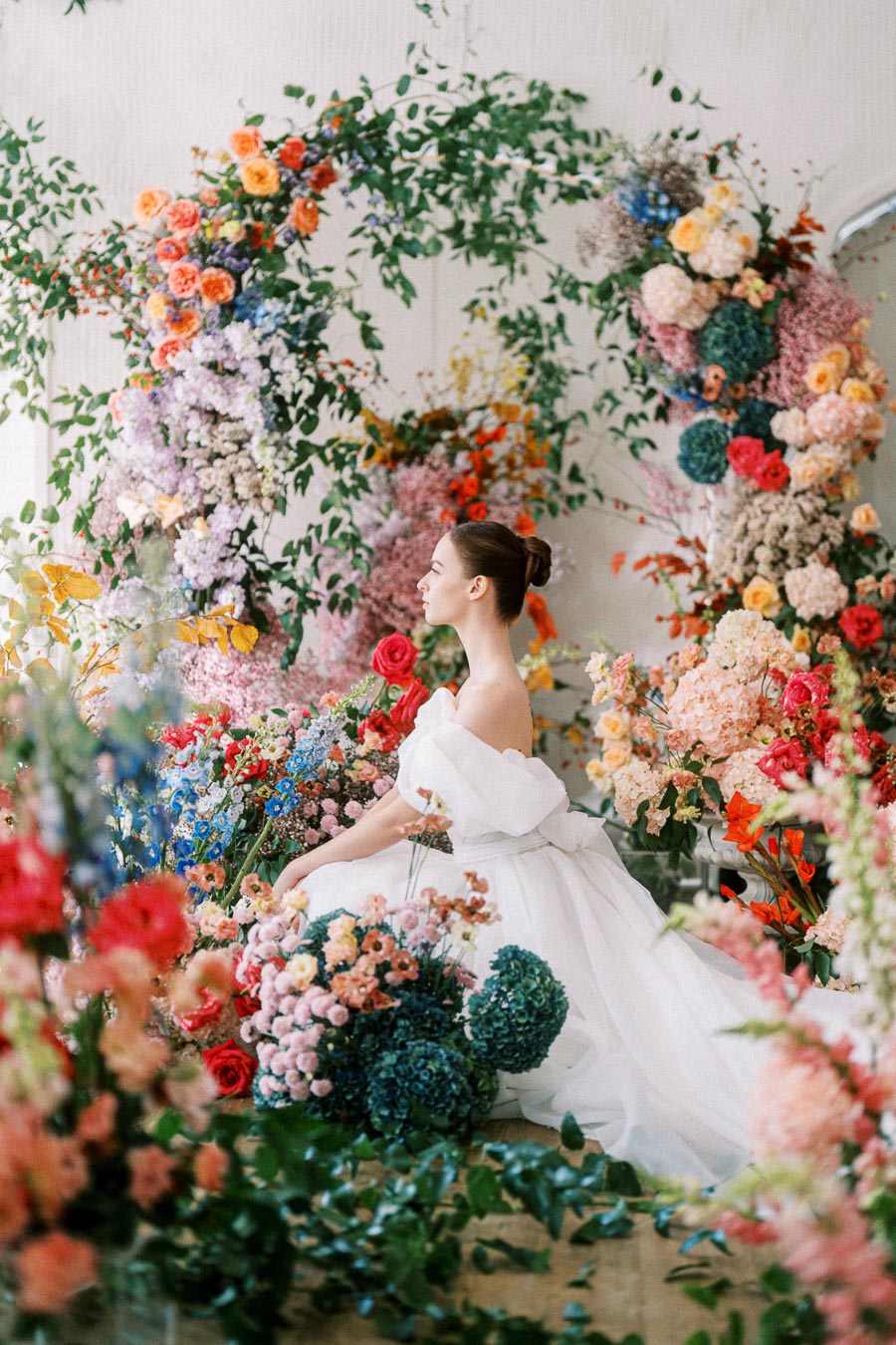 A bride in an elegant white gown sits amidst a vibrant floral arrangement featuring a variety of colorful blooms and lush greenery, creating a romantic garden ambiance.
