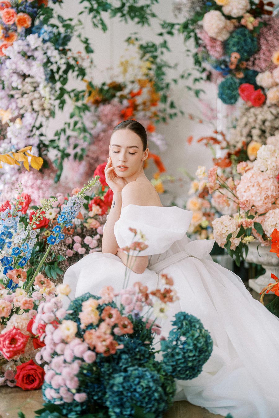 A woman in an elegant white dress poses gracefully among a vibrant array of colorful flowers, including pink, blue, and red blooms, creating a dreamy and romantic setting.