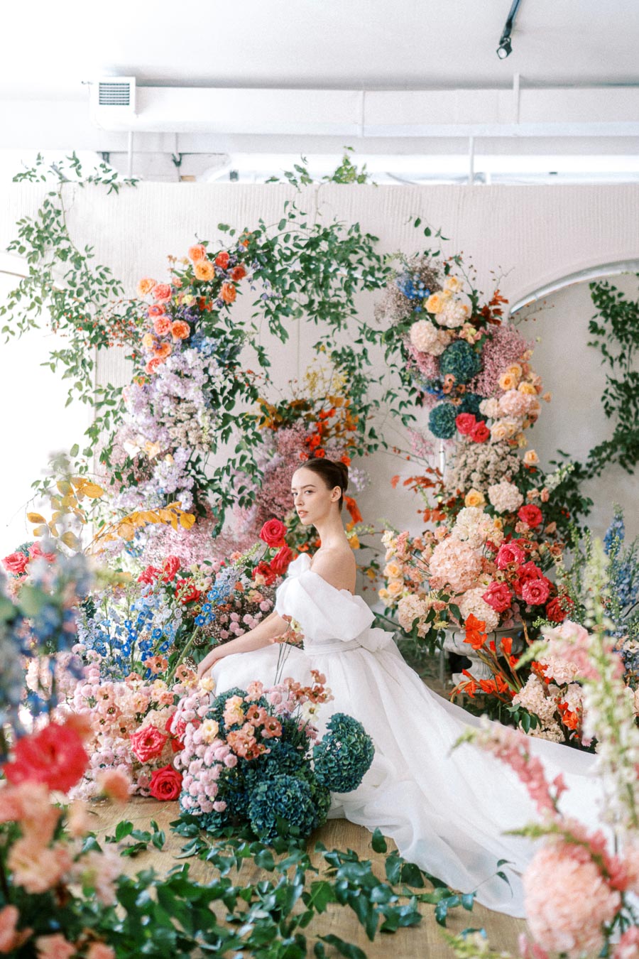 A bride in an elegant white gown surrounded by vibrant, colorful floral arrangements in an indoor setting.
