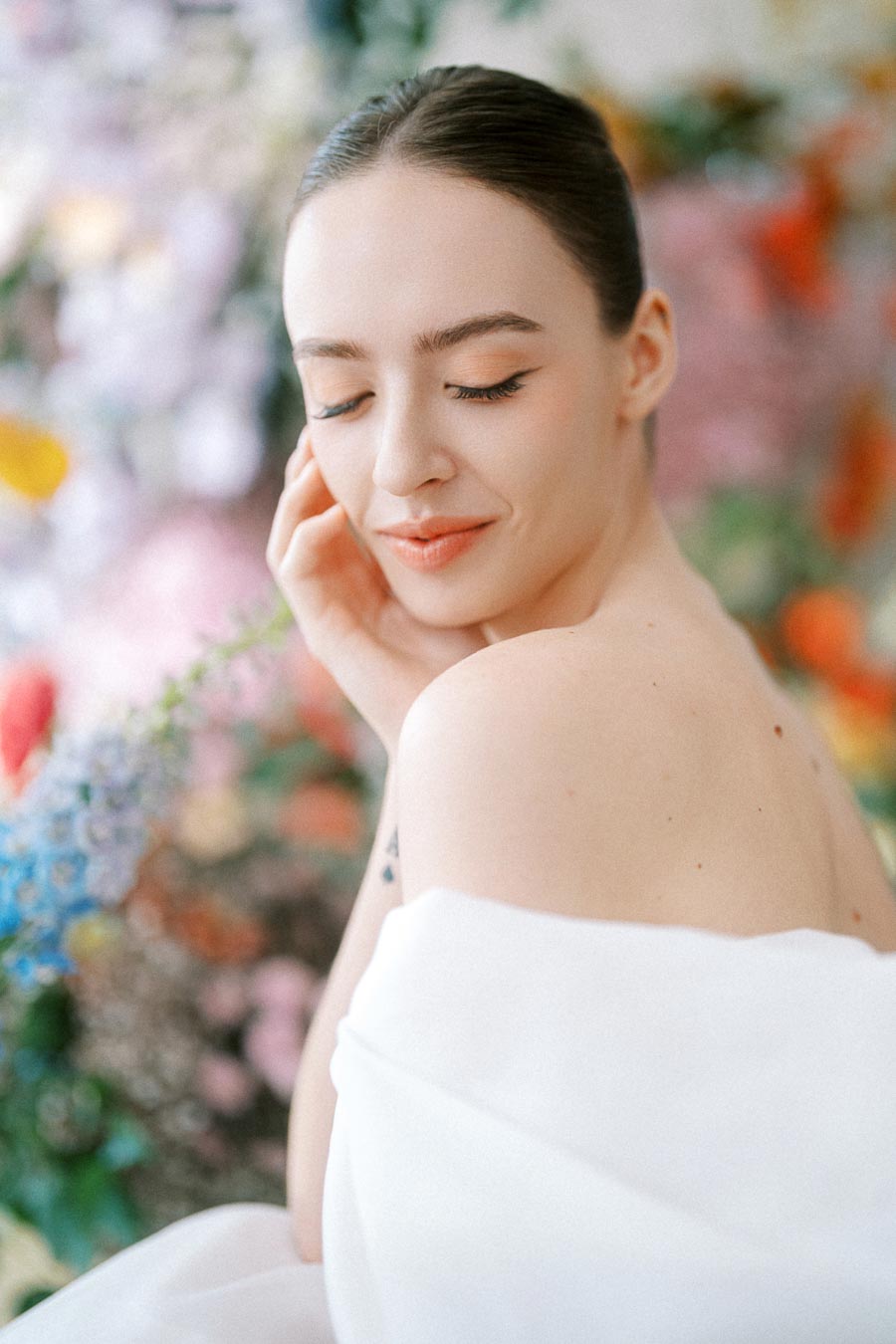 A serene woman wrapped in a white fabric smiling gently with her eyes closed, set against a vibrant floral background.