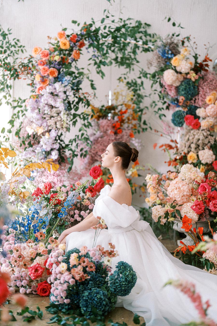 Elegant bride in a flowing white gown surrounded by a vibrant floral arrangement with roses and greenery.