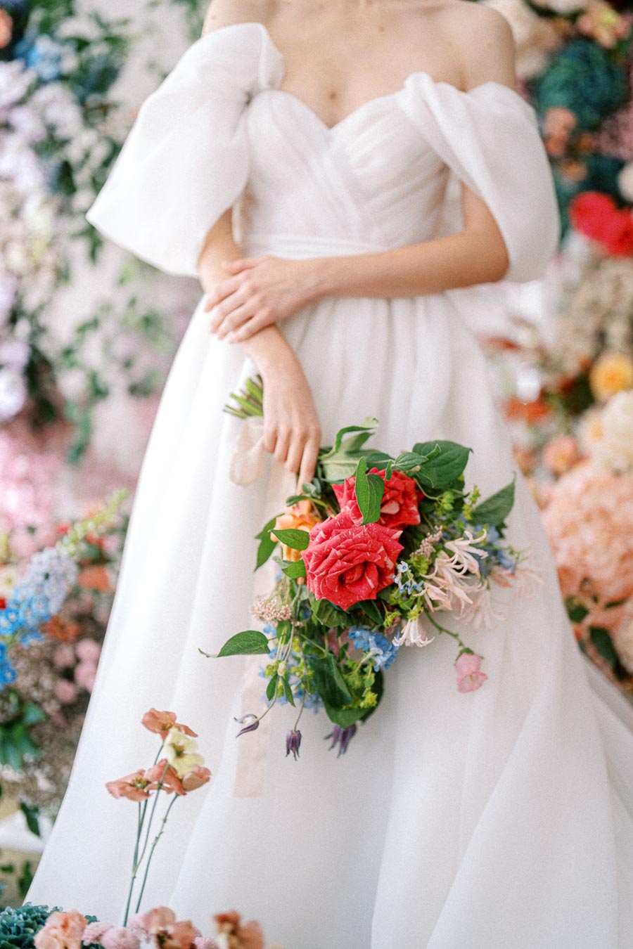 Elegant bride in off-the-shoulder white wedding gown holding a vibrant bouquet of red, pink, and blue flowers amidst a floral backdrop, capturing a romantic wedding theme.