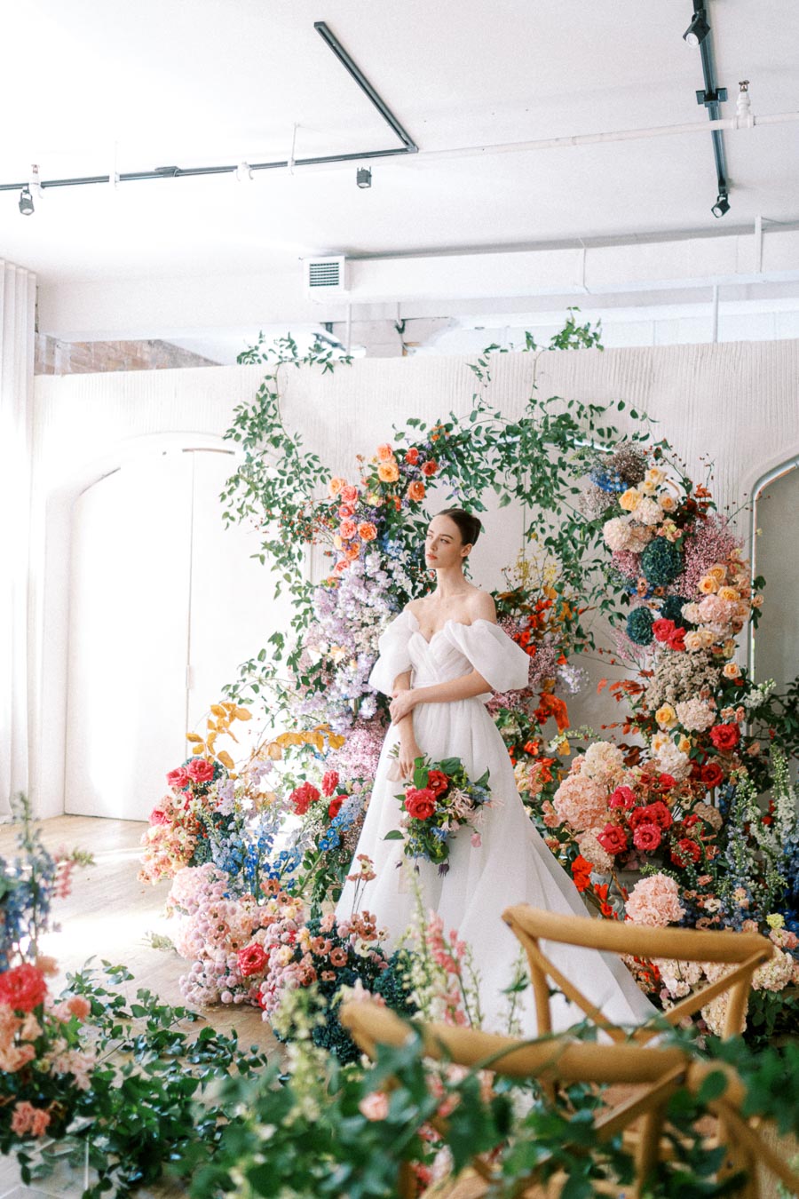 A bride in an elegant white gown stands amidst a vibrant floral backdrop, featuring an array of colorful flowers, creating a romantic wedding setting indoors.