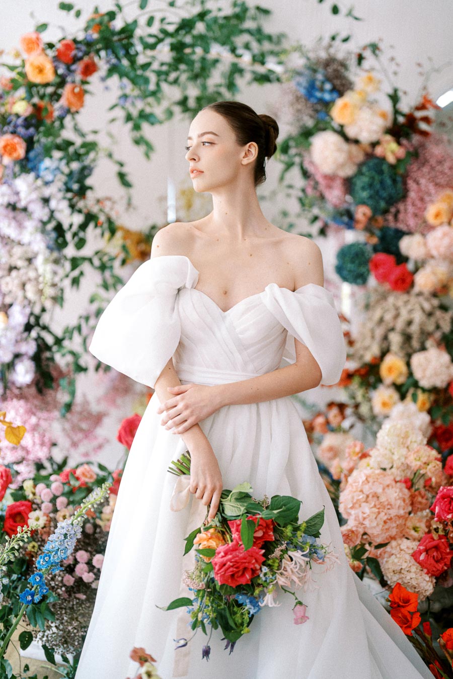 A bride in an elegant off-shoulder white wedding gown holding a vibrant bouquet, surrounded by colorful floral arrangements, looking away with a serene expression.