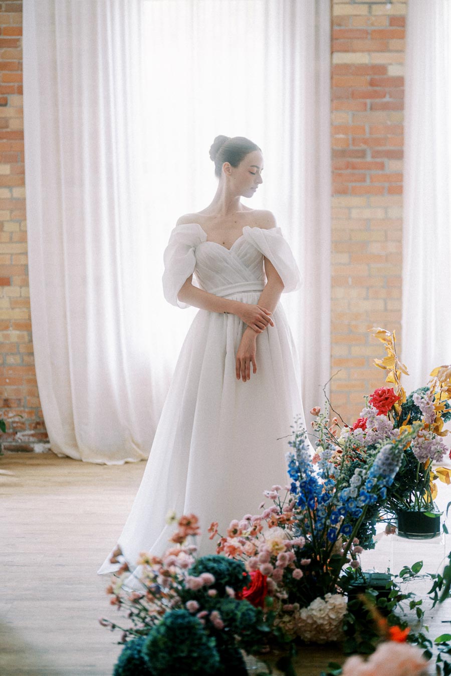 Elegant bride in a flowing white wedding gown surrounded by vibrant floral arrangements in a softly lit room with brick walls and sheer curtains.