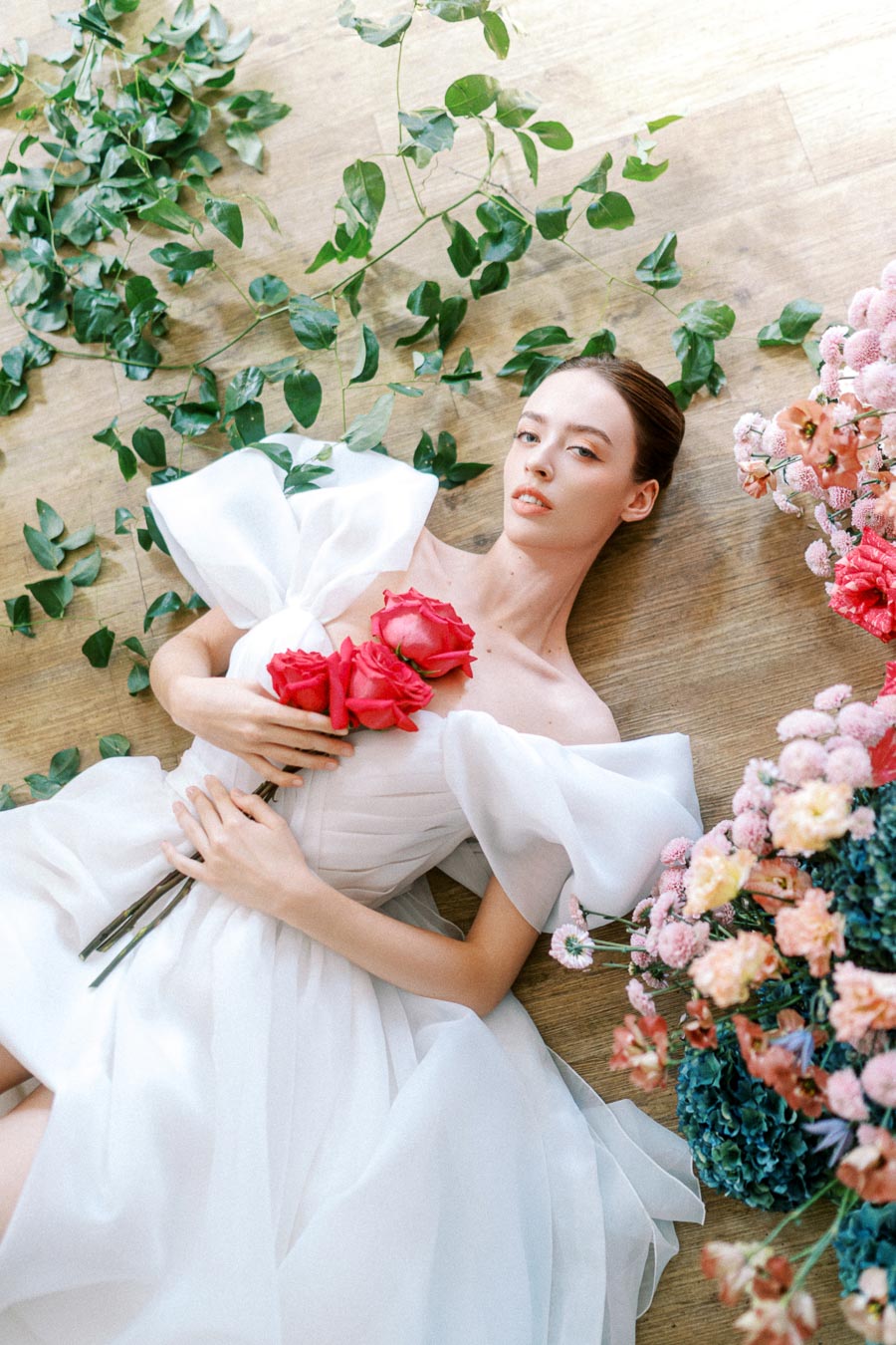Elegant woman in a white dress lying on a wooden floor, holding red roses surrounded by green leaves and vibrant flowers, conveying a serene and romantic atmosphere.