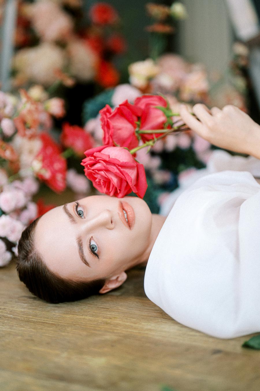 A woman lying down surrounded by vibrant roses, holding pink flowers, with a serene expression, showcasing natural beauty and floral elegance.